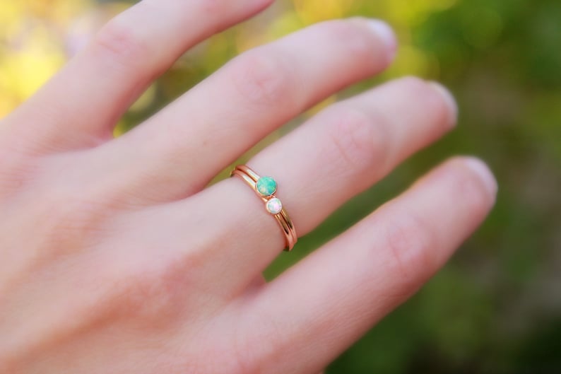 Op de afbeelding: Een ros&eacute;gouden ring met twee kleine edelstenen, een groene en een witte. De ring zit om een vinger, met een vage groene en gele achtergrond.