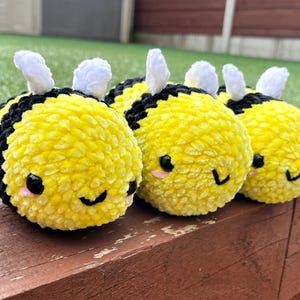May include: Three plush yellow and black bumblebees with white antennae and smiling faces. The bees are made of a soft, textured material and are arranged in a row on a wooden surface. The background includes green grass and a wooden fence.