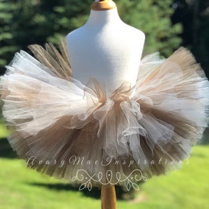 May include: A brown and white tulle tutu on a white mannequin. The tutu is layered and has a ruffled edge. The tutu is on a wooden stand. The text "Avary Mae Inspirations" is visible on the bottom of the image.