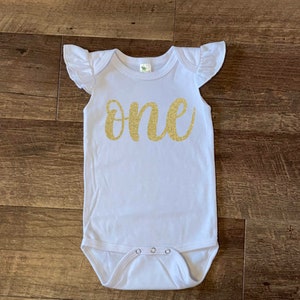 May include: White baby onesie with gold glitter lettering that says "one".