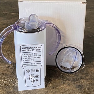 K&ouml;nnte beinhalten: Ein wei&szlig;er Edelstahlbecher mit einem durchsichtigen Deckel und zwei Griffen. Der Becher hat einen Aufkleber mit der Aufschrift "Tumbler Pflegehinweise" und listet auf: Nur Handw&auml;sche, nicht sp&uuml;lmaschinenfest, nicht mikrowellengeeignet und nicht fallen lassen. Der Aufkleber sagt auch "Edelstahl, doppelwandig vakuumversiegelt" und "Hei&szlig;e oder kalte Getr&auml;nke". Der Becher hat einen Aufkleber mit der Aufschrift "Danke".