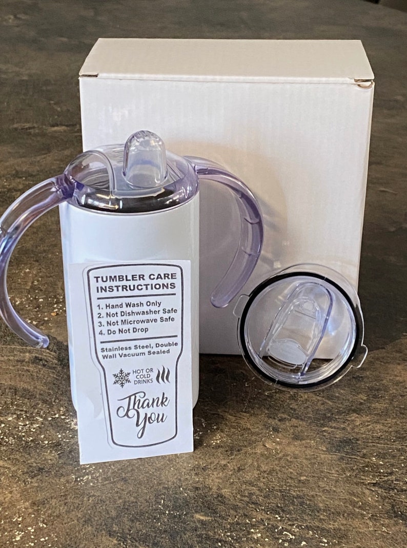 K&ouml;nnte beinhalten: Ein wei&szlig;er Edelstahlbecher mit einem durchsichtigen Deckel und zwei Griffen. Der Becher hat einen Aufkleber mit der Aufschrift "Tumbler Pflegehinweise" und listet auf: Nur Handw&auml;sche, nicht sp&uuml;lmaschinenfest, nicht mikrowellengeeignet und nicht fallen lassen. Der Aufkleber sagt auch "Edelstahl, doppelwandig vakuumversiegelt" und "Hei&szlig;e oder kalte Getr&auml;nke". Der Becher hat einen Aufkleber mit der Aufschrift "Danke".