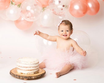 May include: A baby girl in a pink tutu sits in front of a cake with pink and white balloons in the background. The baby is smiling and reaching for the cake.