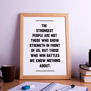 May include: A framed inspirational quote on a wooden desk with a brown coffee mug, a notebook, pens, and a vintage camera. The quote reads: "The strongest people are not those who show strength in front of us, but those who win battles we know nothing about."