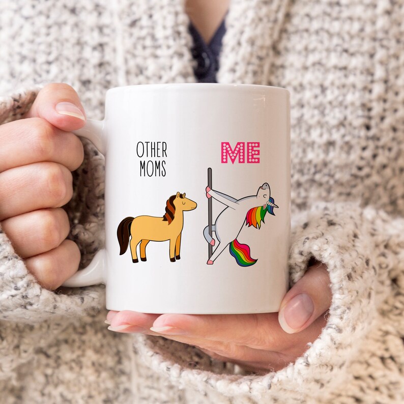 Peut inclure: Un mug en c&eacute;ramique blanche avec une illustration graphique. &Agrave; gauche, un cheval brun avec le texte OTHER MOMS. &Agrave; droite, une licorne blanche &agrave; la crini&egrave;re et &agrave; la queue arc-en-ciel dansant sur une barre avec le texte rose ME au-dessus.