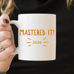 May include: A white ceramic mug featuring the orange text "MASTERED IT!" above the year "2026" with small orange decorative lines surrounding the date. The mug has a smooth finish and a standard handle on the left side for holding.