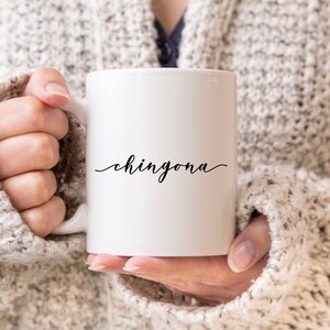 May include: A white ceramic mug featuring the word chingona printed in a black cursive script font on the front. The mug has a smooth, glossy finish and a standard handle on the left side, designed for holding hot or cold beverages in a simple style.