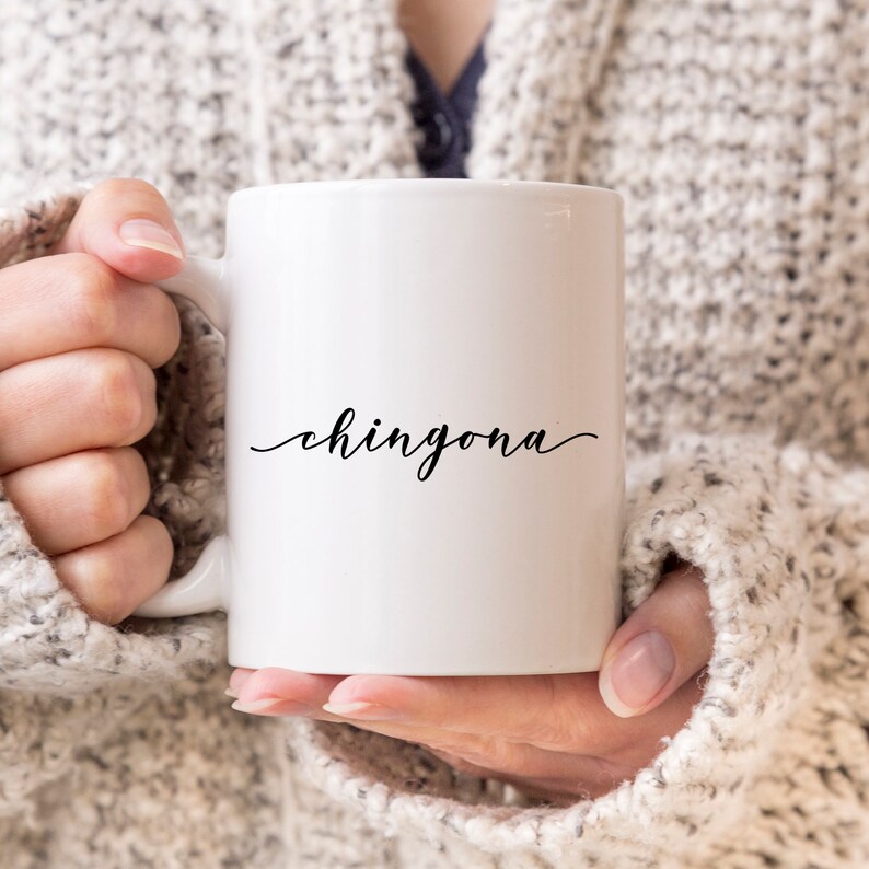 May include: A white ceramic mug featuring the word chingona printed in a black cursive script font on the front. The mug has a smooth, glossy finish and a standard handle on the left side, designed for holding hot or cold beverages in a simple style.