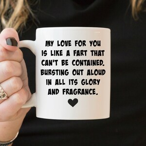 May include: A white ceramic mug featuring black text that reads: MY LOVE FOR YOU IS LIKE A FART THAT CAN'T BE CONTAINED. BURSTING OUT ALOUD IN ALL ITS GLORY AND FRAGRANCE. A small black heart icon is printed directly below the text on the mug.