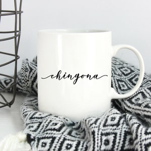 May include: A white ceramic mug featuring the word chingona printed in a black cursive script font on the front. The mug has a standard cylindrical shape with a curved handle on the right side and a smooth, glossy finish throughout the exterior.