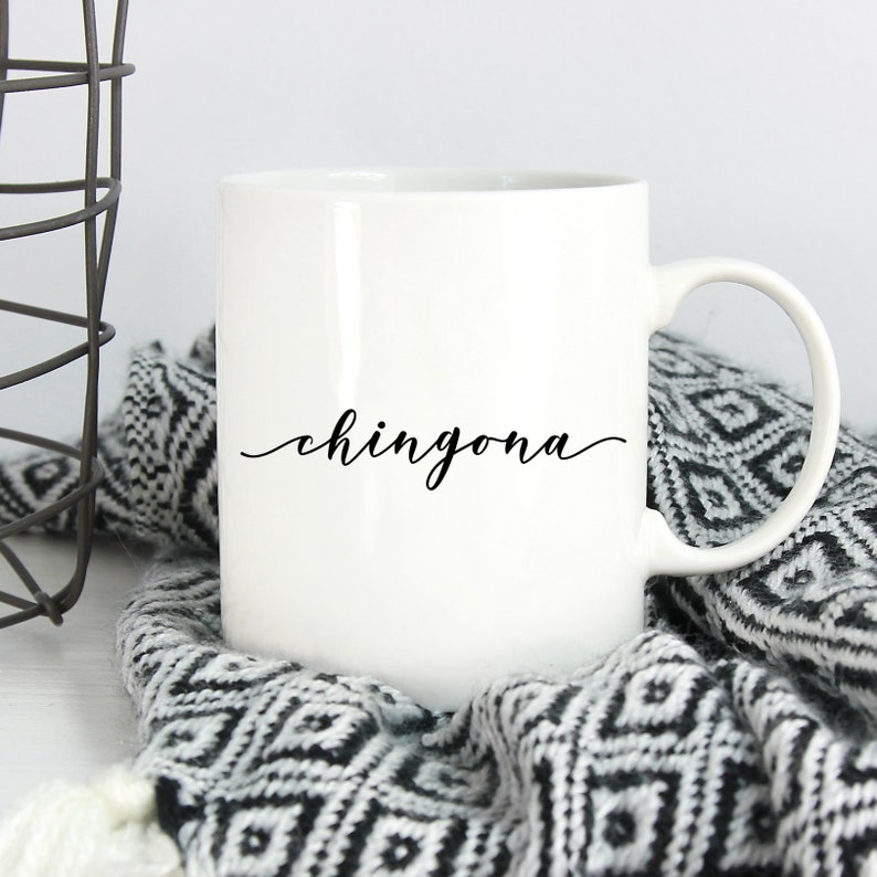 May include: A white ceramic mug featuring the word chingona printed in a black cursive script font on the front. The mug has a standard cylindrical shape with a curved handle on the right side and a smooth, glossy finish throughout the exterior.