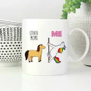 Peut inclure: Un mug en c&eacute;ramique blanc avec une illustration graphique. &Agrave; gauche, un cheval beige &agrave; crini&egrave;re brune est accompagn&eacute; du texte "OTHER MOMS". &Agrave; droite, une licorne blanche &agrave; crini&egrave;re et queue arc-en-ciel danse sur une barre, avec le texte "ME" en rose.