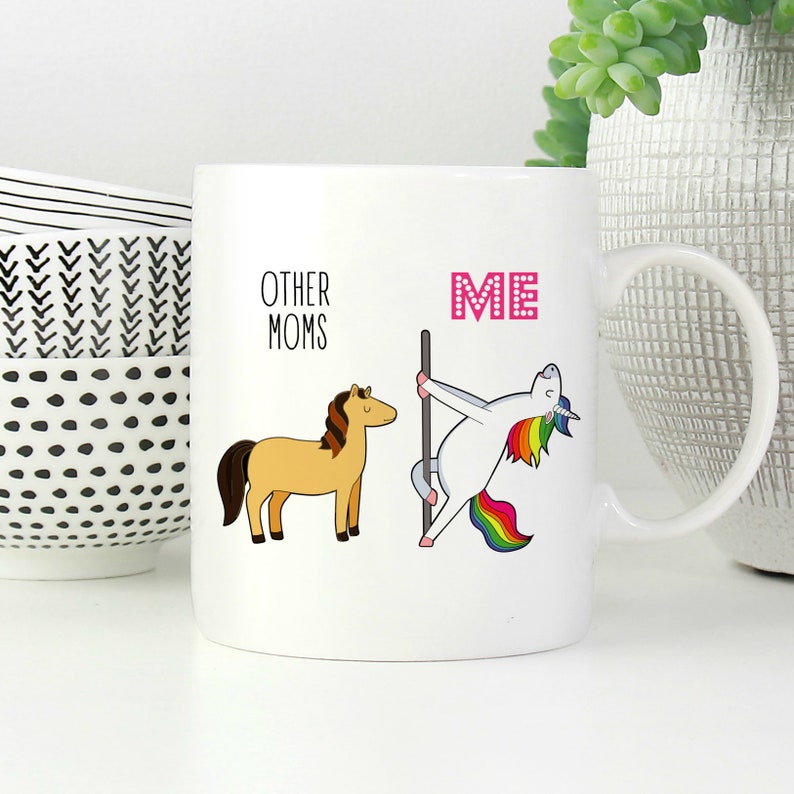 Peut inclure: Un mug en c&eacute;ramique blanc avec une illustration graphique. &Agrave; gauche, un cheval beige &agrave; crini&egrave;re brune est accompagn&eacute; du texte "OTHER MOMS". &Agrave; droite, une licorne blanche &agrave; crini&egrave;re et queue arc-en-ciel danse sur une barre, avec le texte "ME" en rose.
