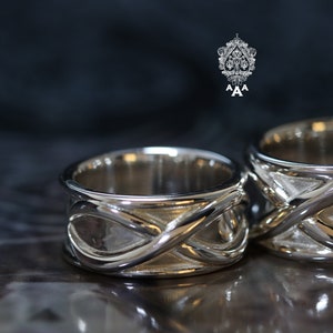 Könnte beinhalten: Zwei silberne Eheringe mit einem komplizierten, ineinander verschlungenen Design. Die Ringe sind poliert und haben eine glatte Oberfläche.