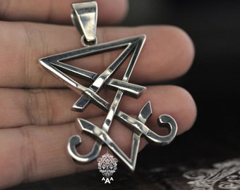 Può includere: Un ciondolo d'argento con un design geometrico stilizzato a forma triangolare e linee intricate. Il ciondolo è su una mano.