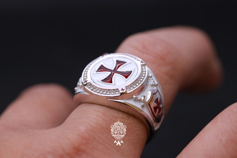 May include: A silver ring with a white enamel center featuring a red cross. The ring has a shield design on the side with a red cross.