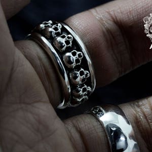 May include: A silver ring featuring a band of skulls. The ring is worn on a person's finger.