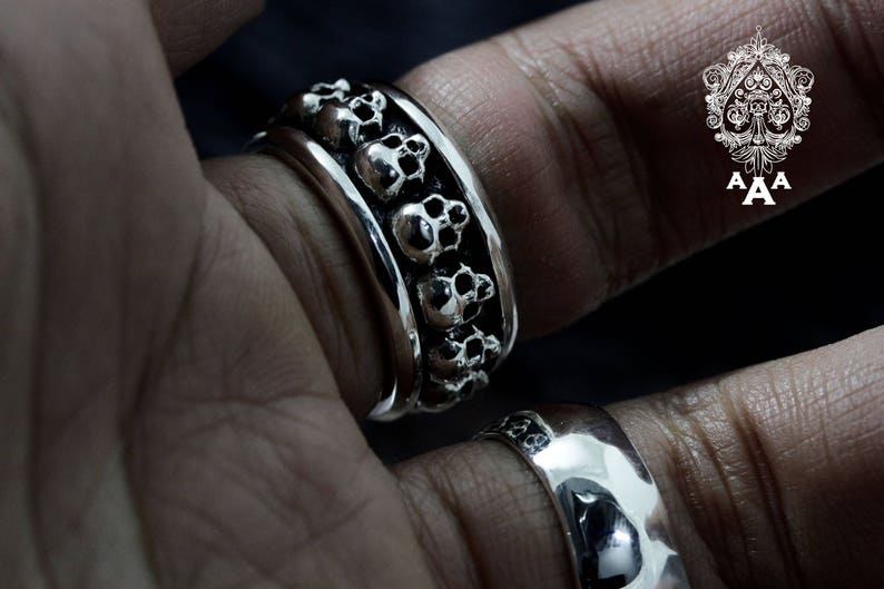 May include: A silver ring featuring a band of skulls. The ring is worn on a person's finger.