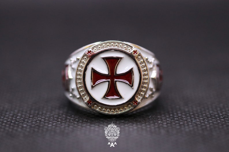 May include: A silver ring with a white enamel circle featuring a red cross. The ring has a textured band and small red accents.