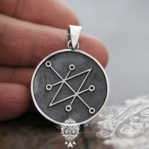 Pode incluir: Um pingente de prata com fundo preto e um design geométrico prateado. O design apresenta uma forma de diamante com linhas que se estendem de cada canto até um círculo.