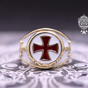 May include: A gold ring featuring a white enamel band and a red cross design. The cross is set within a gold circle, with decorative gold detailing. The ring has the text "AAA" and a decorative emblem.