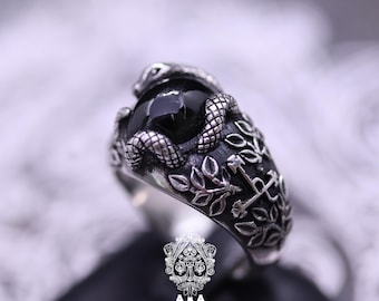 May include: A silver ring featuring a black gemstone encircled by a detailed snake design. The ring's band is adorned with leaf and anchor motifs, creating a gothic or vintage aesthetic. The ring is displayed against a blurred background.