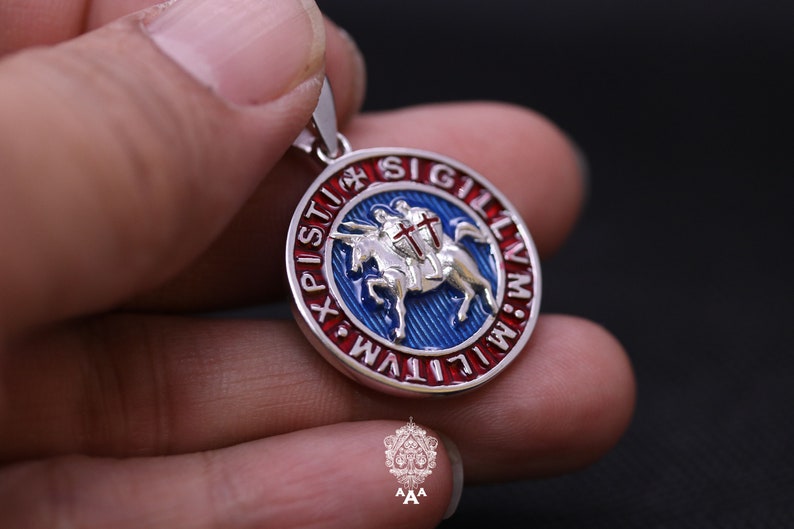 May include: A silver pendant with a red, white, and blue enamel design. The pendant features a knight riding a horse with a cross on his shield. The text around the edge reads "SIGILLVM MILITVM XPI STV", which is Latin for "Seal of the Knights of Christ".