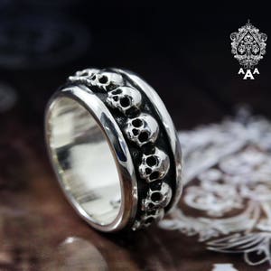 May include: A silver ring with a band of skulls around the circumference. The skulls are detailed and have a slightly raised design.