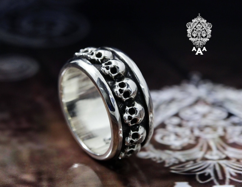 May include: A silver ring with a band of skulls around the circumference. The skulls are detailed and have a slightly raised design.