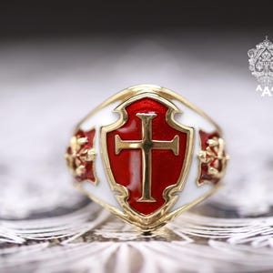 Könnte beinhalten: Goldfarbener Ring mit rotem und weißem Emaille-Design. Der Ring hat eine Schildform mit einem goldenen Kreuz in der Mitte. Der Ring hat ein detailliertes Blumenmuster an den Seiten.