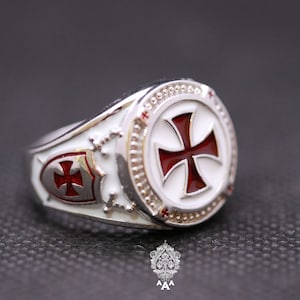 May include: A silver ring with a white enamel background and a red enamel cross. The ring has a shield design with a smaller red cross on the side.