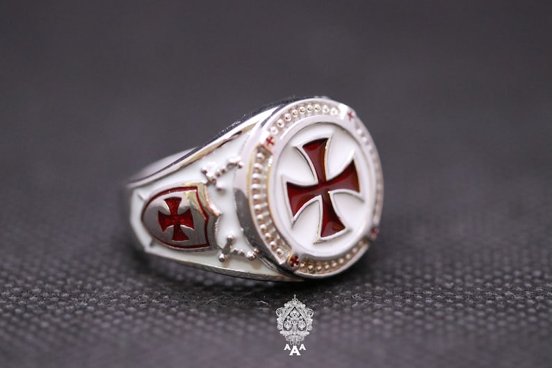 May include: A silver ring with a white enamel background and a red enamel cross. The ring has a shield design with a smaller red cross on the side.