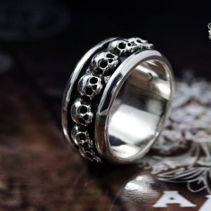 May include: A silver ring with a band of skulls around the circumference. The ring is on a dark brown surface.