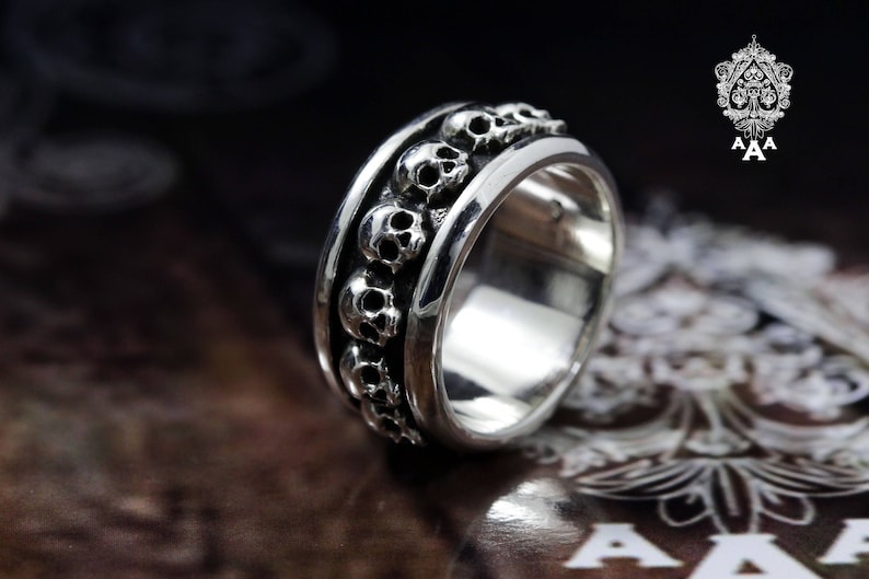 May include: A silver ring with a band of skulls around the circumference. The ring is on a dark brown surface.