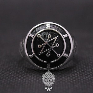 May include: A silver ring with a black enamel inlay featuring a pentagram with a circle around it. The ring has a silver band with engraved symbols.