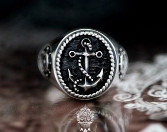 Puede incluir: Un anillo de plata con un diseño de ancla negra. El ancla está rodeada por un borde de cuerda y el anillo tiene una superficie texturizada.