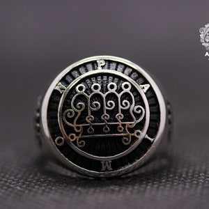 以下が含まれることがあります： 黒のエナメルの中央に、詳細で複雑なデザインが施された銀の指輪。指輪の縁には「M P A M」の文字が刻まれている。