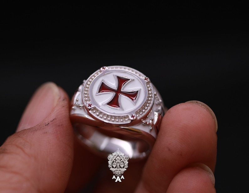 May include: A silver ring with a white enamel cross with a red center. The cross is surrounded by a circle of small white dots.