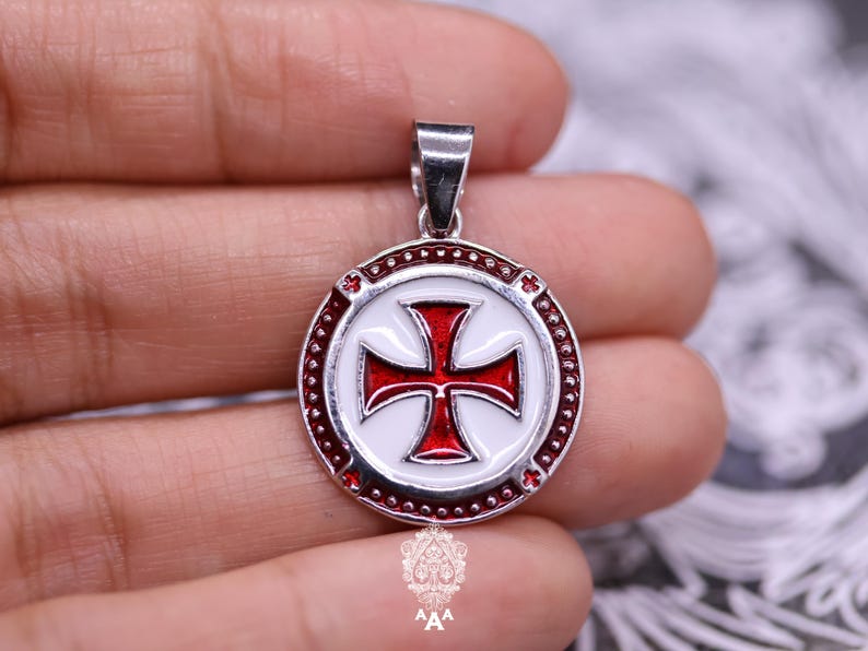 以下が含まれることがあります： 赤と白のマルタ十字のデザインのシルバーペンダント。十字は白い背景に配置され、小さな赤いアクセントが付いた赤い縁で囲まれています。ペンダントにはチェーン用のループが付いています。