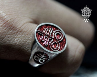 May include: A silver ring with a red enamel design featuring a stylized symbol. The symbol is a combination of a cross and a circle.