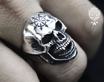 May include: A silver skull ring with a detailed design. The skull has a large, open mouth and a starburst design on the forehead.
