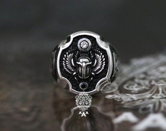 May include: A silver ring featuring a black background and an intricate design of a scarab beetle with wings. A single clear stone is set in the centre of the ring.