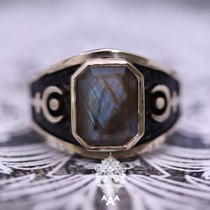 Könnte beinhalten: Ein goldfarbener Ring mit einem großen, rechteckigen, schillernden Edelstein. Das Band ist schwarz mit einem sich wiederholenden Halbmond-Design. Der Edelstein hat blaue und braune Farbtöne.
