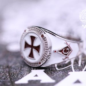 May include: A silver ring with a white enamel design featuring a red cross and a Masonic symbol. The ring has a textured band.