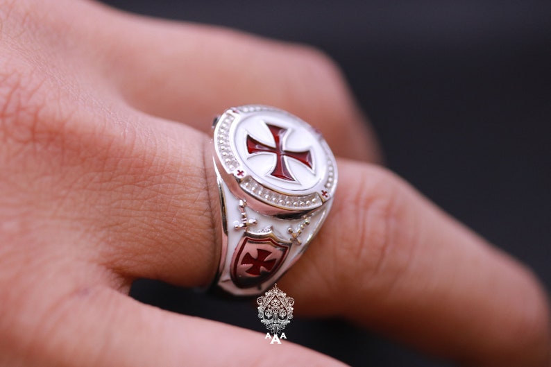 May include: A silver ring with a white enamel cross and a red enamel cross on a shield. The ring is worn on a finger.