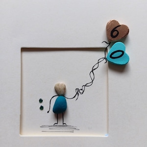 Peut inclure: Une &oelig;uvre d'art en galets bleus et blancs repr&eacute;sentant une personne tenant une cha&icirc;ne de ballons en forme de c&oelig;ur bleus et bruns avec les chiffres 6 et 0.