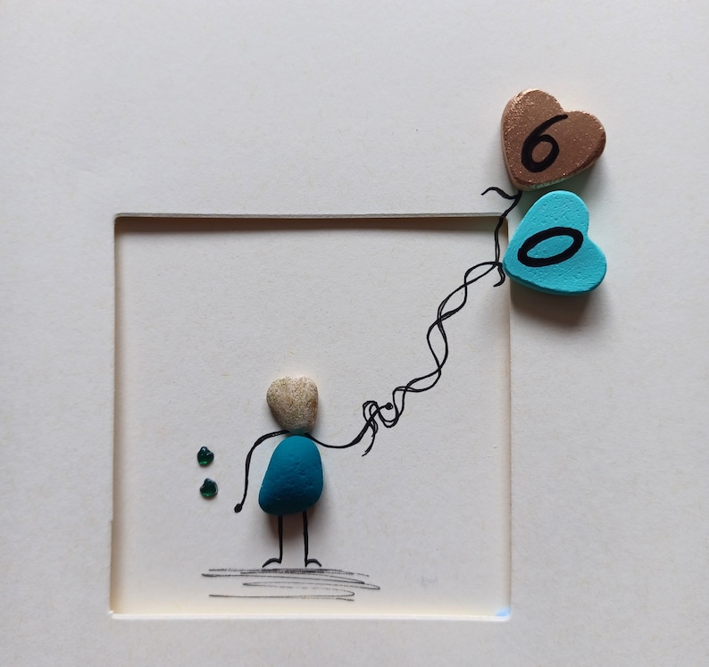 Peut inclure: Une &oelig;uvre d'art en galets bleus et blancs repr&eacute;sentant une personne tenant une cha&icirc;ne de ballons en forme de c&oelig;ur bleus et bruns avec les chiffres 6 et 0.