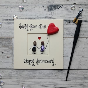 May include: A framed anniversary artwork with the text "Forty years of us" and "Happy Anniversary". The artwork features two pebble figures holding hands, a red heart balloon, and a small red heart. A black calligraphy pen is on the side.