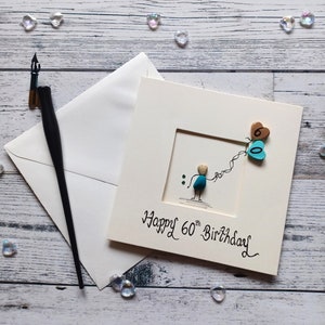 Peut inclure: Une carte blanche avec une illustration dessin&eacute;e &agrave; la main d'une personne tenant deux ballons en forme de c&oelig;ur. Les ballons sont d&eacute;cor&eacute;s des chiffres "60" et le texte "Happy 60th Birthday" est &eacute;crit sous l'illustration.