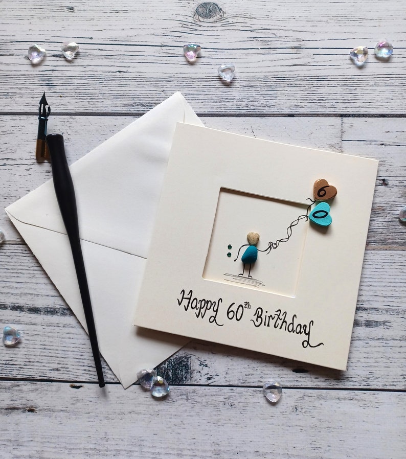 Peut inclure: Une carte blanche avec une illustration dessin&eacute;e &agrave; la main d'une personne tenant deux ballons en forme de c&oelig;ur. Les ballons sont d&eacute;cor&eacute;s des chiffres "60" et le texte "Happy 60th Birthday" est &eacute;crit sous l'illustration.